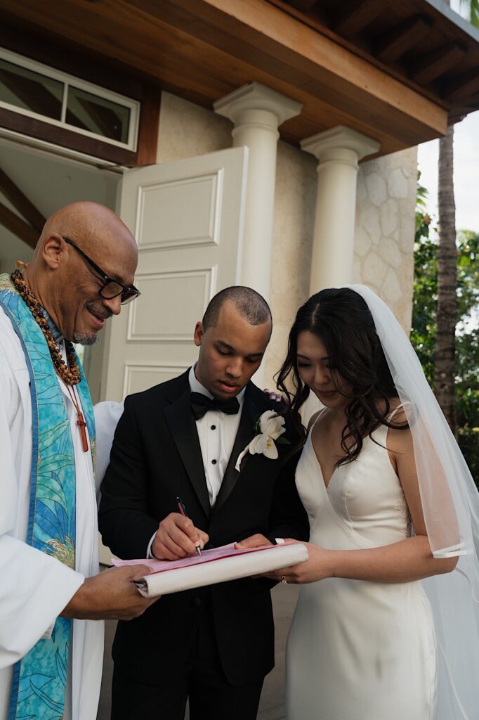 Bride and Groom sign their marriage certificate with the officiant outside beautiful chapel at Four Seasons Oahu in Hawaii. 