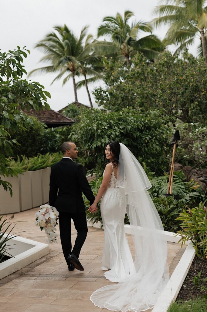 Bride looks behind her as her and the groom walk down brick path surrounded by tropical plants and Palm trees outside Four Seasons Oahu in Hawaii.