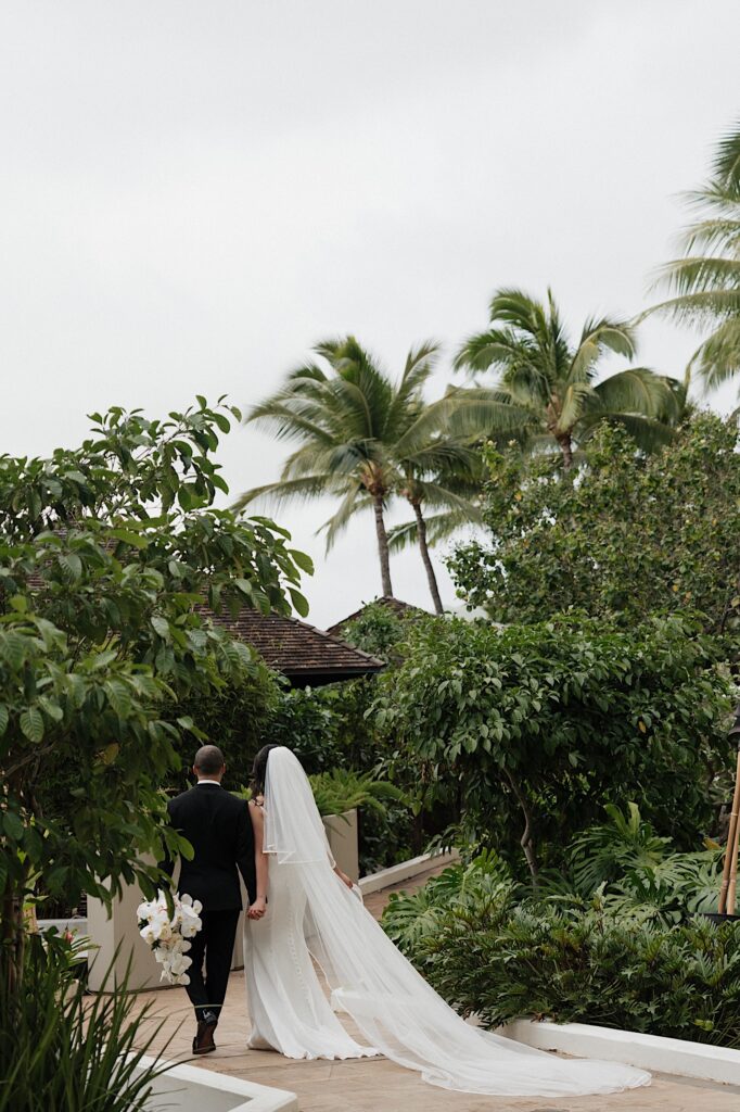 Bride and groom walk together holding hands down brick path surrounded by tropical plants and Palm trees outside Four Seasons Oahu in Hawaii.