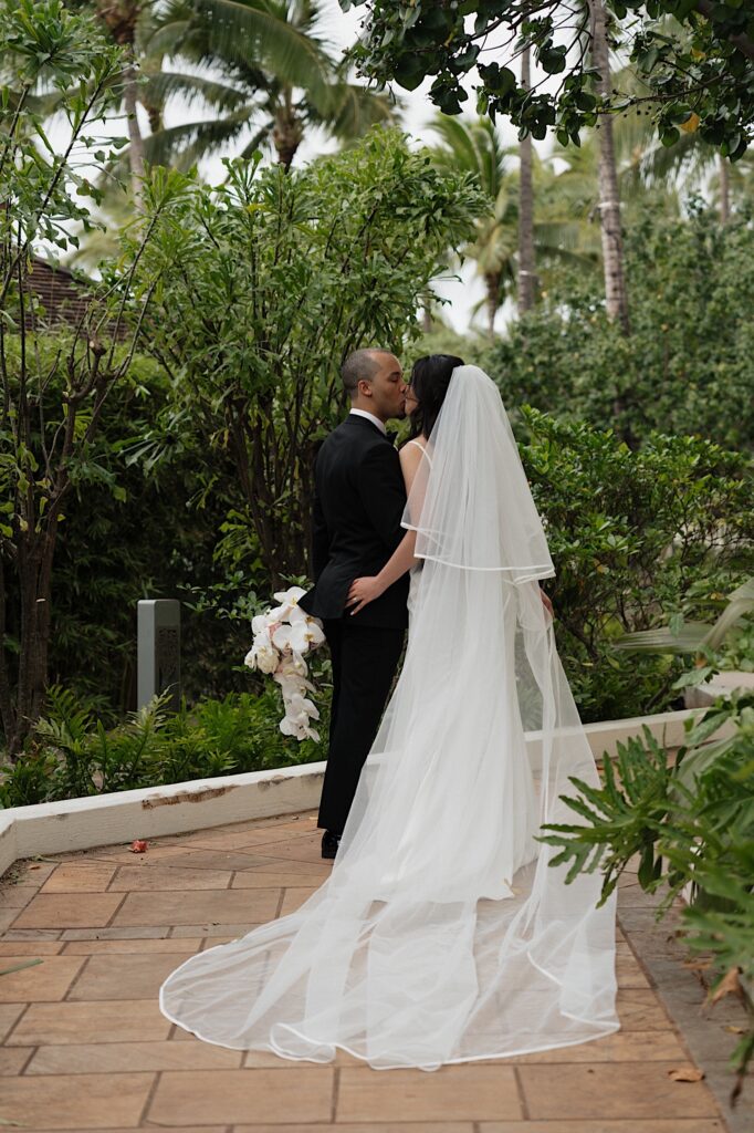 Bride and groom with their backs turned share a kiss in front of beautiful tropical plants and flowers outside Four Seasons Oahu in Hawaii. 