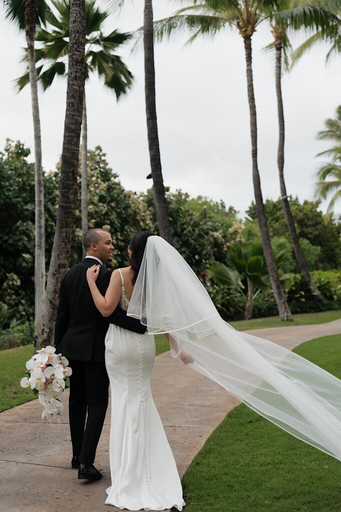 Bride in black tux holds bride's tropical flower bouquet and hold's brides waist as they look at each other and walk down brick path outside Four Seasons Oahu in Hawaii. 