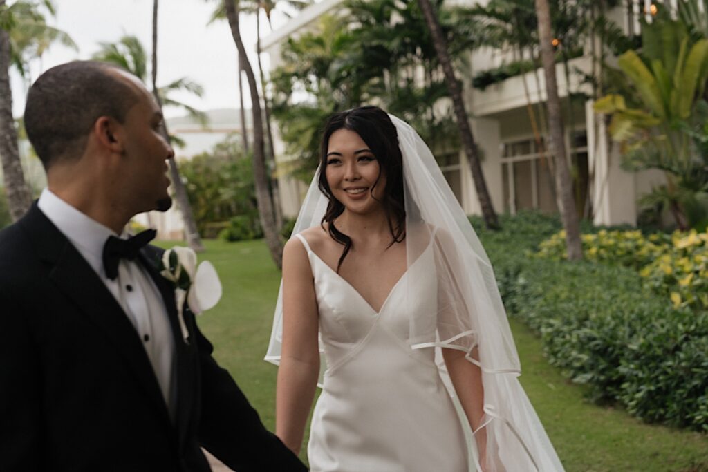 Groom in black tux looks at Bride in beautiful white dress and long white veil as they walk through the grass outside Four Seasons Oahu in Hawaii. 