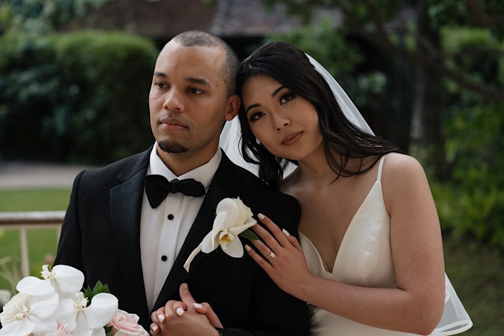 Bride in white gown rests her head against the Grooms as they pose for intimate wedding portraits outside Four Seasons Oahu in Hawaii. 