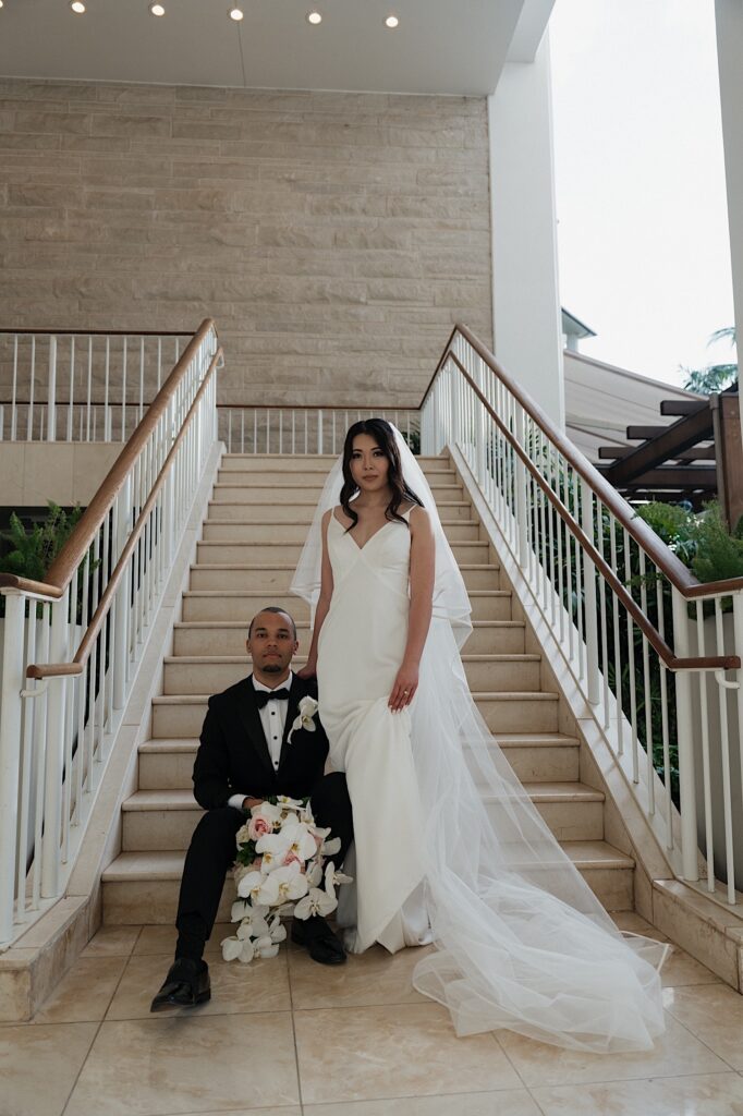 Groom sits on the bottom of the stairs holding Bride's bouquet next to the Bride standing as they pose for wedding portraits at the Four Seasons Oahu in Hawaii. 