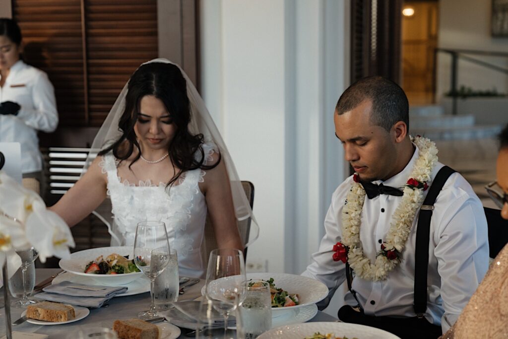 Bride in short white dress and Groom with lei eat salad at their wedding reception at the Four Seassons Oahu in Hawaii. 