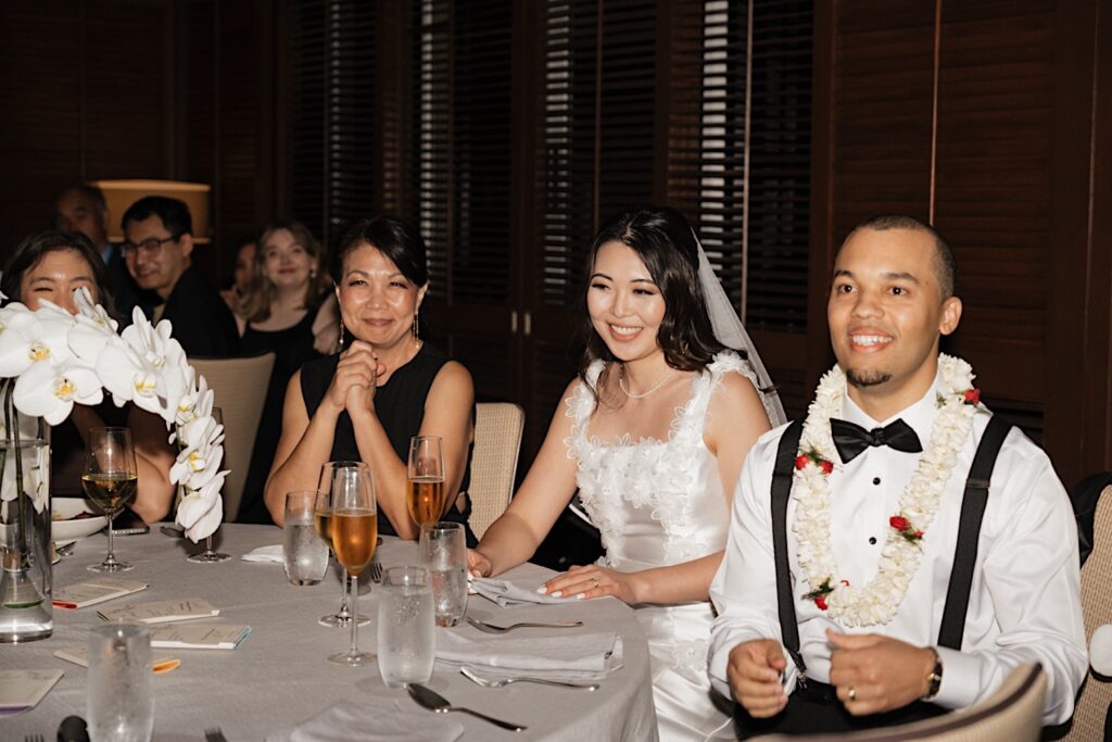 Groom in lei sits next to Bride and bride's mother during wedding speeches at intimate wedding reception at the Four Seasons Oahu in Hawaii. 