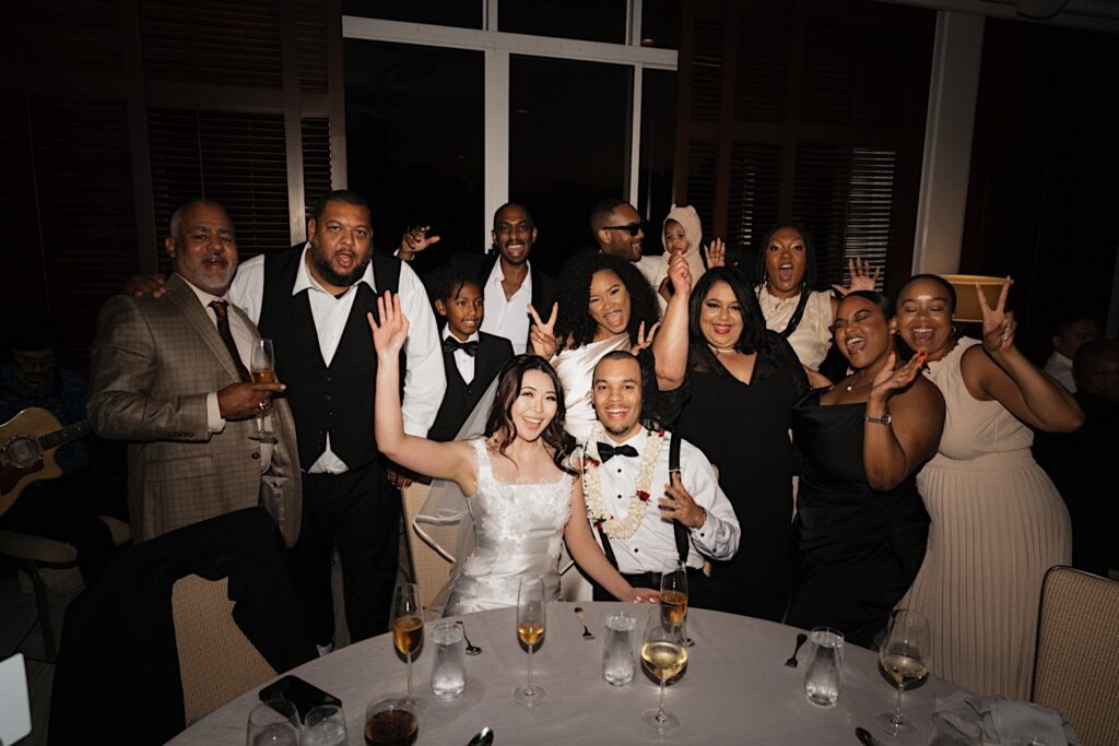 Bride and Groom sit at table as their family surrounds them posing with peace fingers during wedding reception at Four Seasons Oahu in Hawaii. 