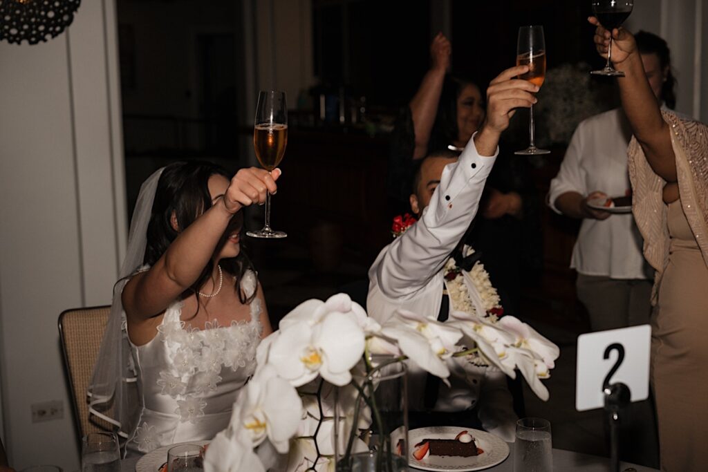 Bride and Groom raise champagne glasses for toast sitting at table during reception at Four Seasons Oahu in Hawaii. 