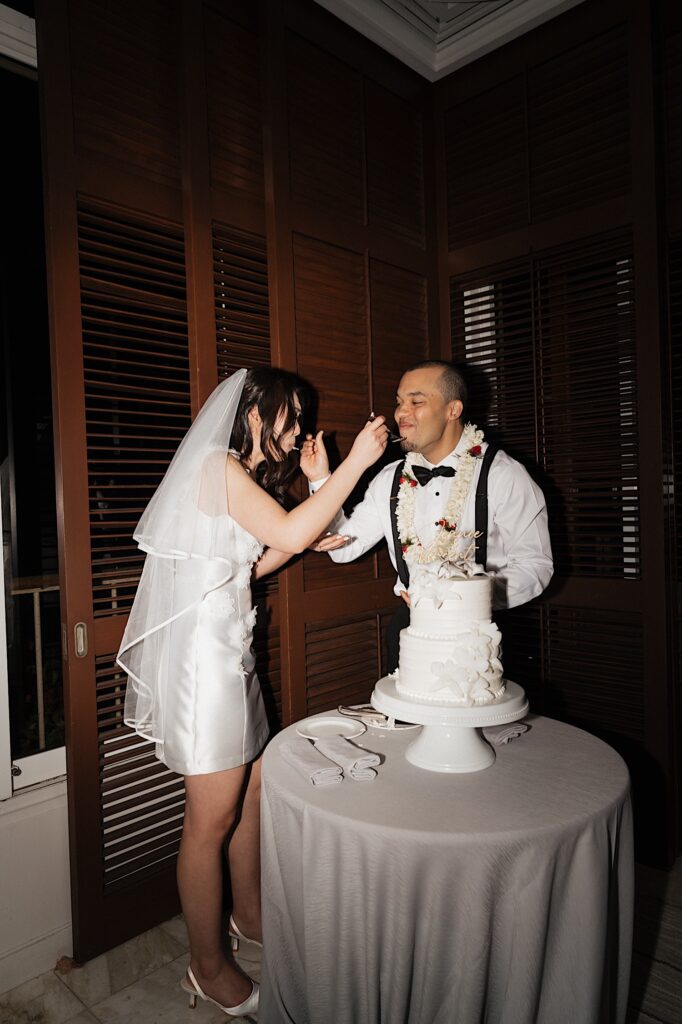 Bride in short white reception dress and Groom in Lei stand next to cake table feed each other cake at wedding reception at Four Season Oahu in Hawaii.