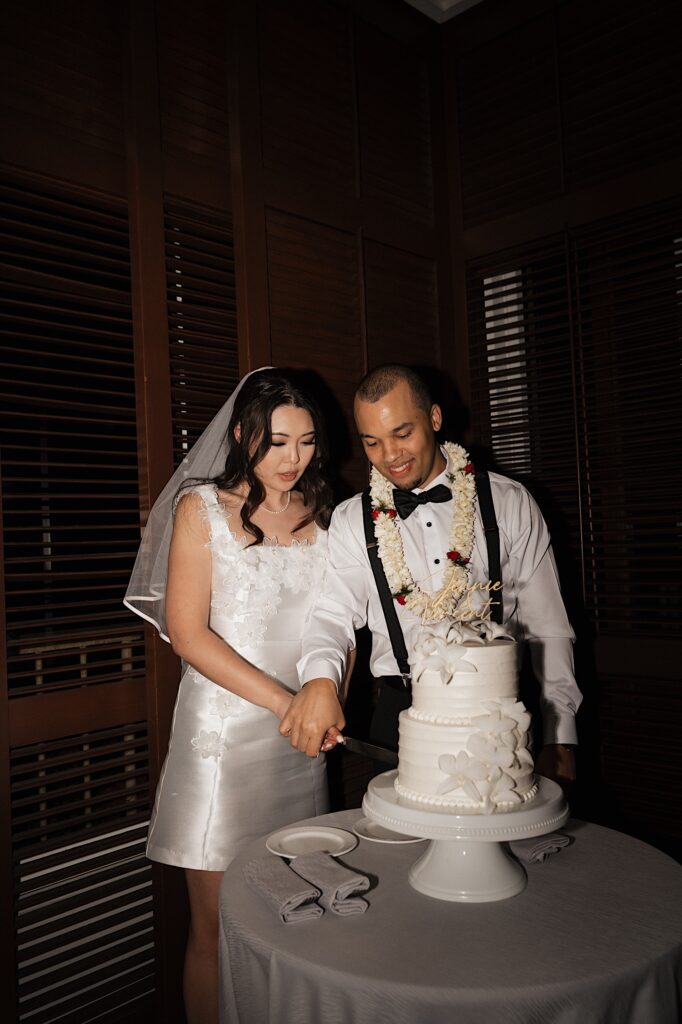 Bride in short white reception dress and Groom in Lei stand next to cake table cutting cake at wedding reception at Four Season Oahu in Hawaii.