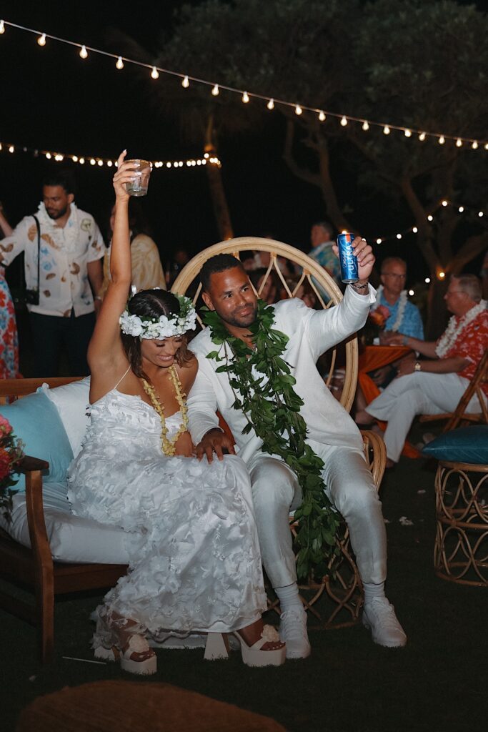 Bride and Groom sit on wicker thrones and raise their drinks in the air as they celebrate during their wedding reception at Turtle Bay in Oahu Hawaii. 