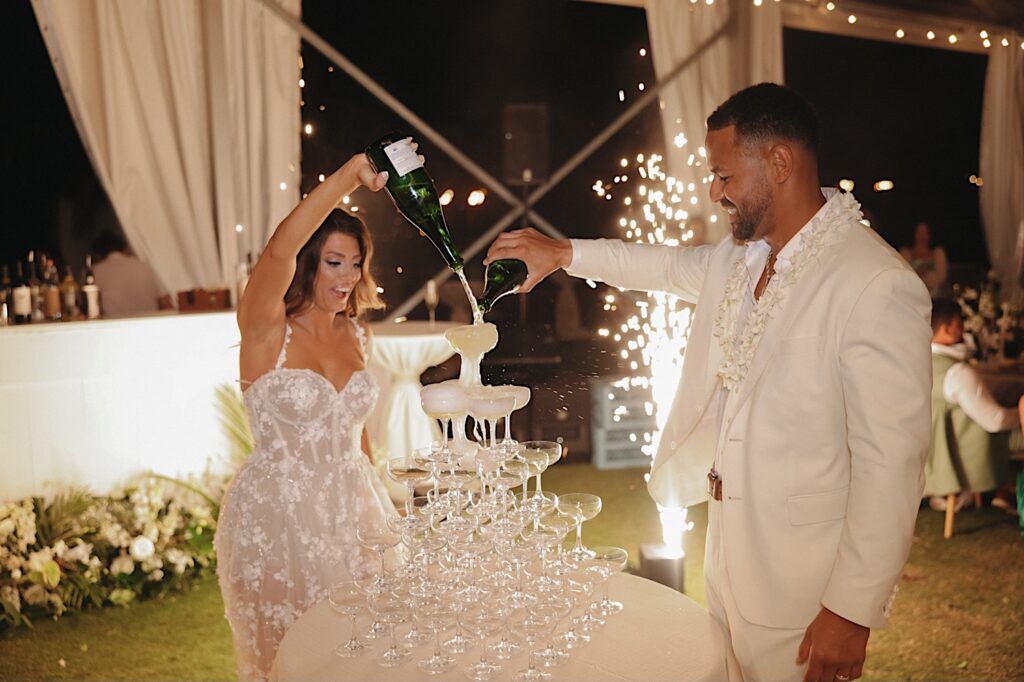 Bride and Groom pour champagne on champagne tower under lit clear tent during wedding reception at Turtle Bay wedding venue in Oahu Hawaii 