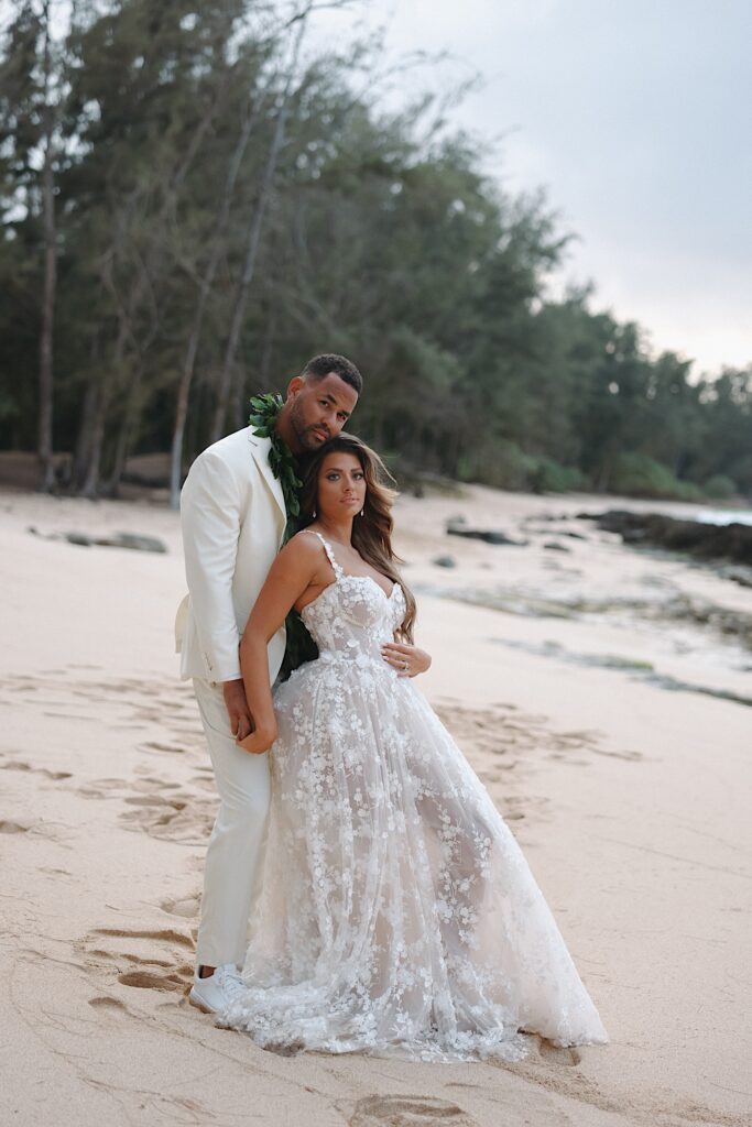 Bride in floral decal dress and Groom in white tux stand on Turtle Bay beach surrounded by trees in Oahu Hawaii. 