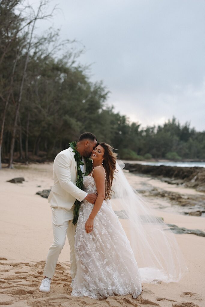 Bride in floral decal dress and Groom in white tux kiss on Turtle Bay beach surrounded by trees in Oahu Hawaii. 