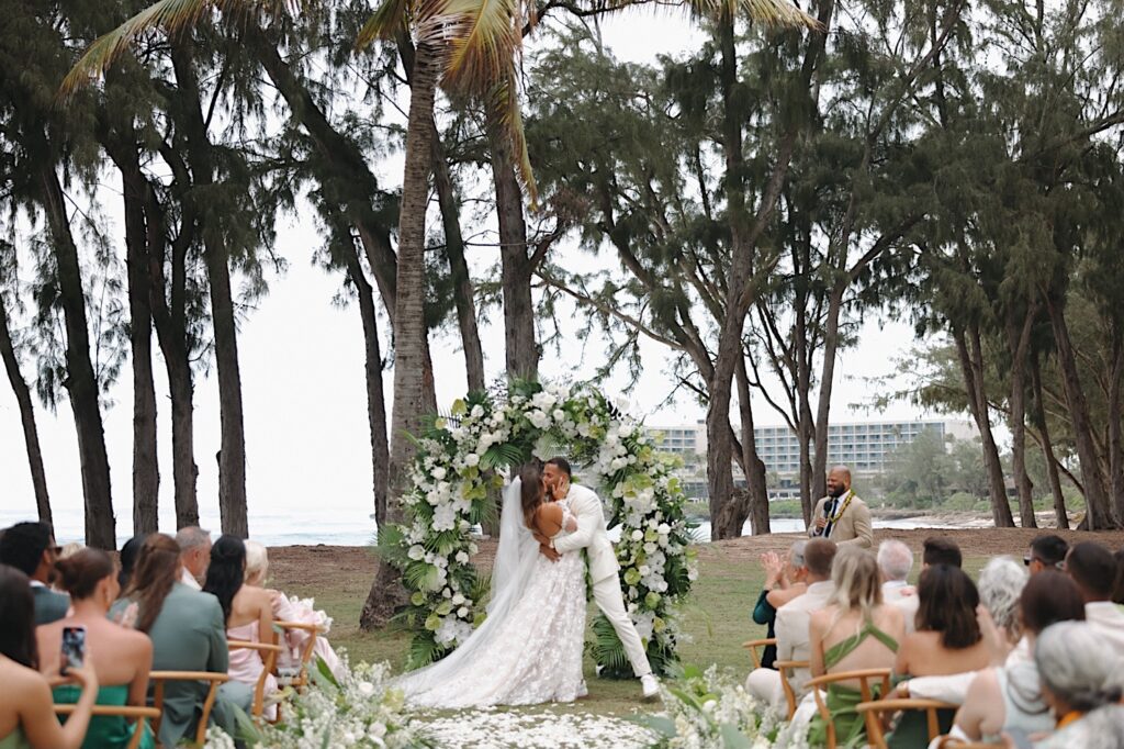 Bride and Groom kiss in front of flower arch covered in white flowers and greenery with Turtle Bay Hotel in the background. 