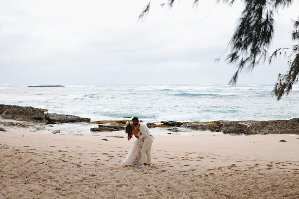 Groom in white tux dips bride for a kiss on the beach overlooking ocean in Oahu Hawaii.