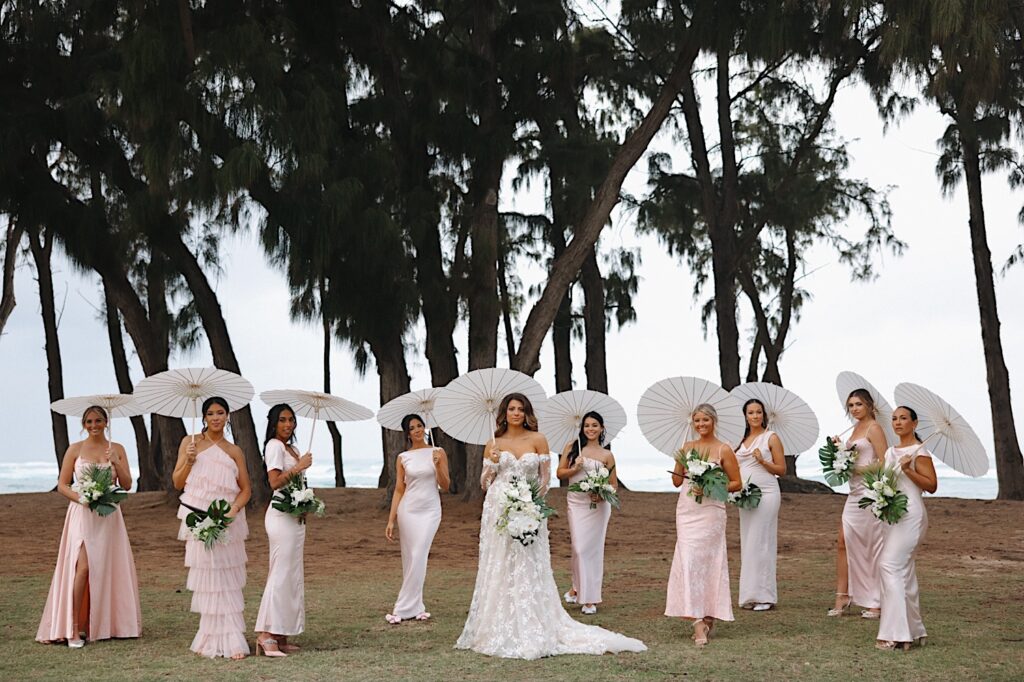 Bride in floral dress is surrounded with bridesmaids and blush dresses all carrying white parasol umbrellas with the ocean in the background in Oahu Hawaii.  