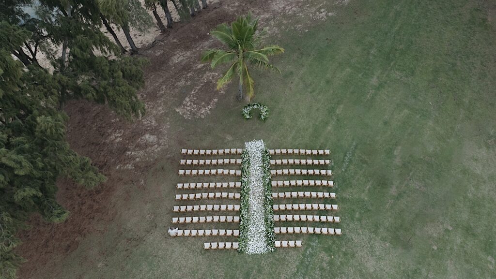 Shot from above, chairs are lined up for the ceremony with white flowers lining the aisle and a flower arch and the end of the aisle. The set-up is surrounded by palm trees near the beach at Turtle Bay in Oahu, Hawaii.