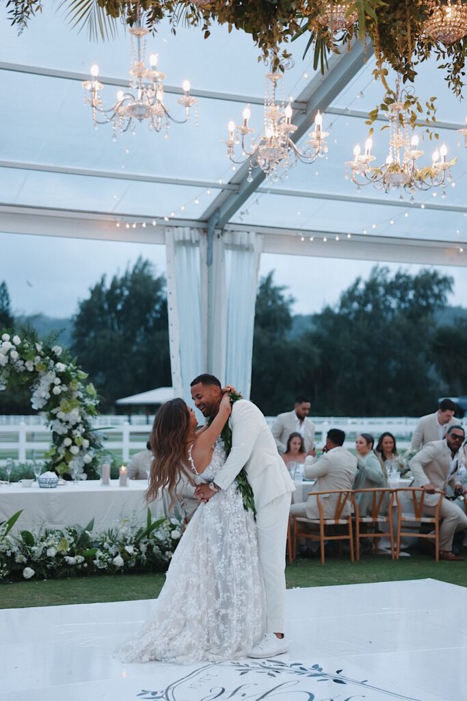 Bride in flower dress dances with Groom in white tux underneath clear tent with chandeliers hanging from the ceiling during wedding reception at Turtle Bay Wedding venue. 