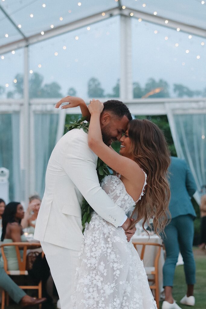 Bride in flower dress dances with Groom in white tux underneath clear tent with chandeliers hanging from the ceiling during wedding reception at Turtle Bay Wedding venue. 