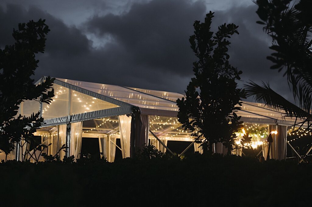 Clear Tent is lit up with dark clouds in the sky surrounded by palm trees.