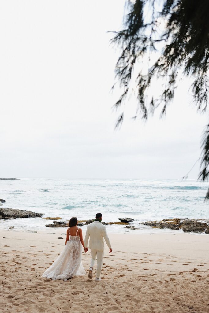 Bride in flower dress and groom in white tux walk along beach with their backs towards the camera and the ocean in the background at Turtle Bay in Oahu Hawaii. 