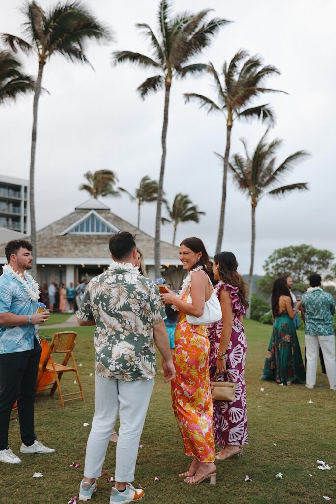 Guests in bright floral Hawaiian shirts and floral dresses stand outside of Turtle Bay Wedding venue socializing during cocktail hour. 