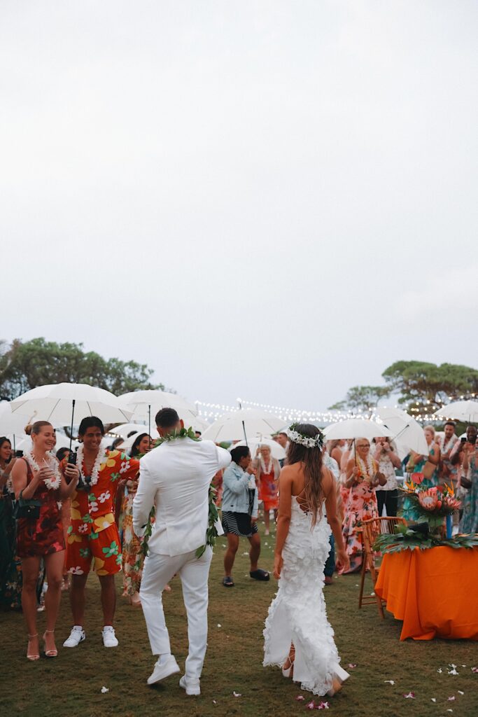 Bride in floral dress and Groom in white tux enter reception where guests in bright floral patterns are holding white umbrellas. 