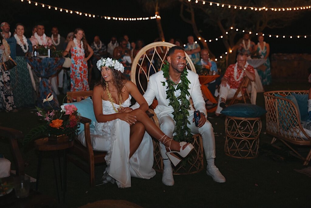 Bride and Groom sit on wicker thrones and smile at their guests as they celebrate during their wedding reception at Turtle Bay in Oahu Hawaii. 