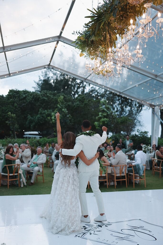 Bride and Groom raise their fists in the air in celebration under clear tent at their wedding reception at Turtle Bay in Oahu, Hawaii.