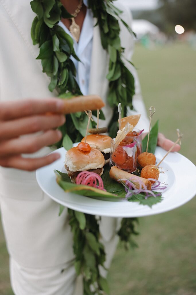 Man holds a plate of delicious cocktail food including sliders and wraps sitting on tropical leaves during cocktail hour at Turtle Bay in Oahu, Hawaii.