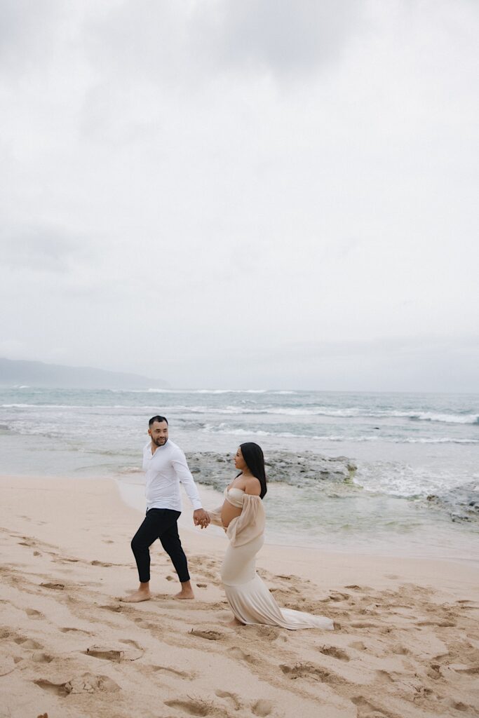 Woman with pregnant belly holds hands with her partner while walking along the beach with ocean in the background in Oahu, Hawaii. 