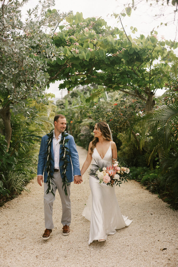 Bride and Groom look at each other as they walk on a sanded path with tropical foliage around them.