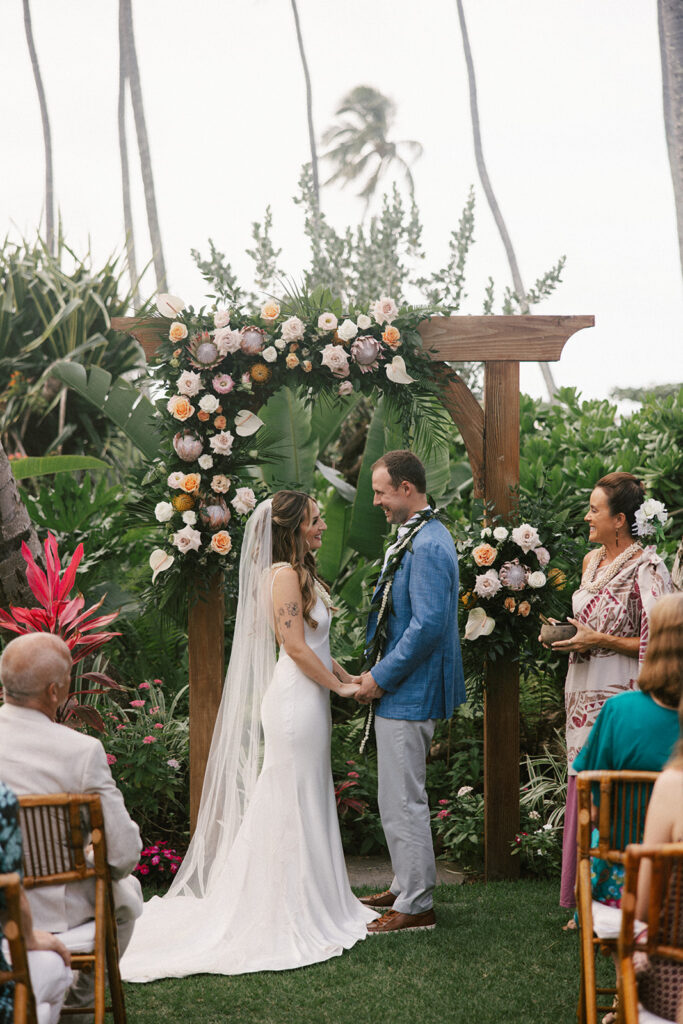 Groom smiles widely as bride looks at him. They stand in front of a large wood arch during their wedding ceremony and their officiant stands of to the side. 