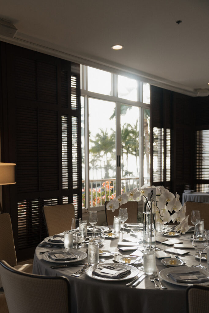 Hawaii wedding photographer captures a wedding reception table with tropical center pieces. The table sits near a window overlooking palm trees and the ocean. 