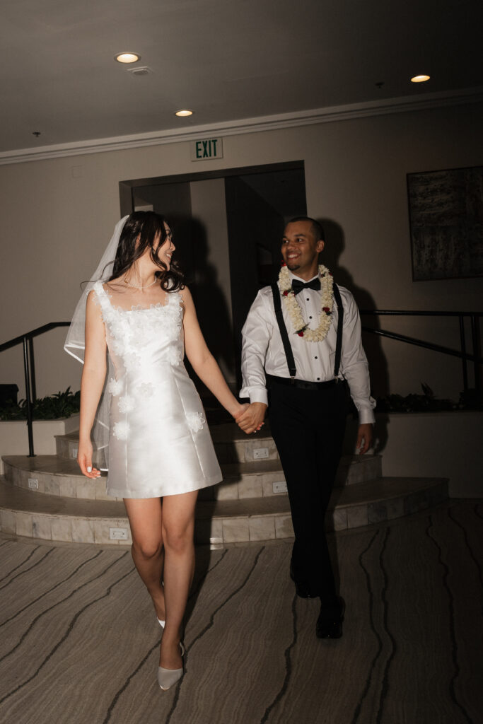 Bride in a short white reception dress looks back at groom as they hold hands and walk into a reception room in Hawaii. 