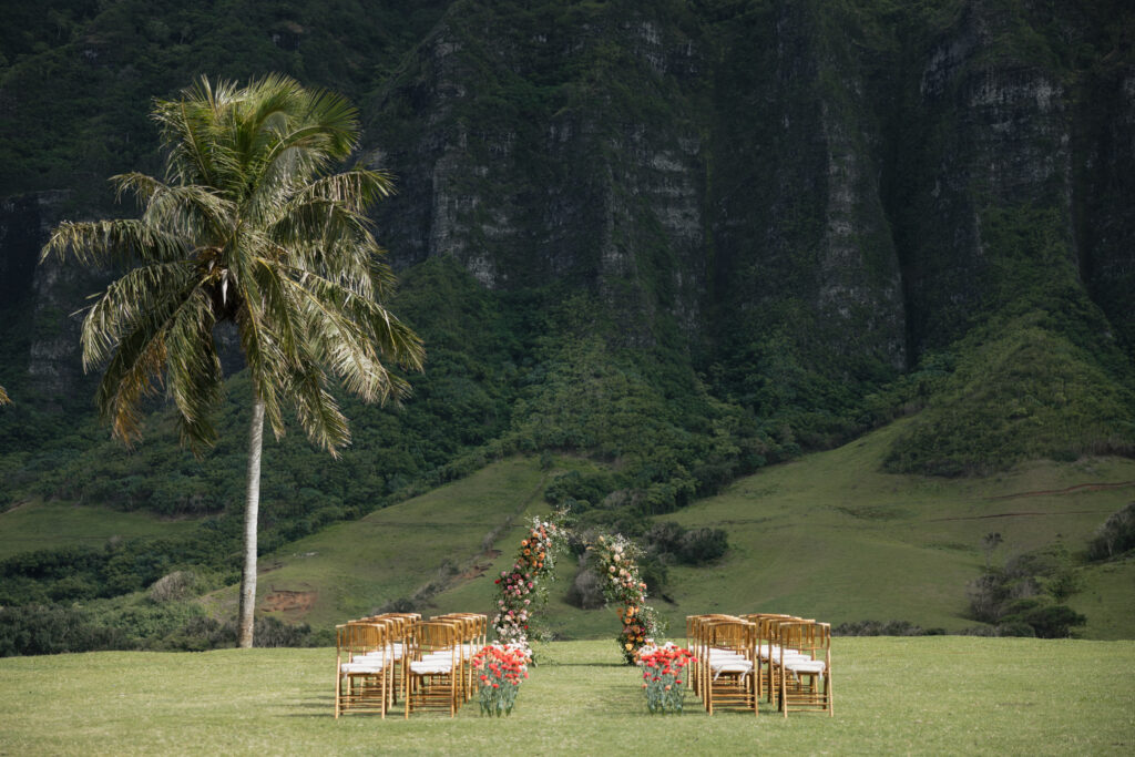 Wooden chairs are set up in the middle of a grassy area with a large flower bouquet center in the aisle. Behind is a large cliff in Hawaii with a palm tree beside it. 