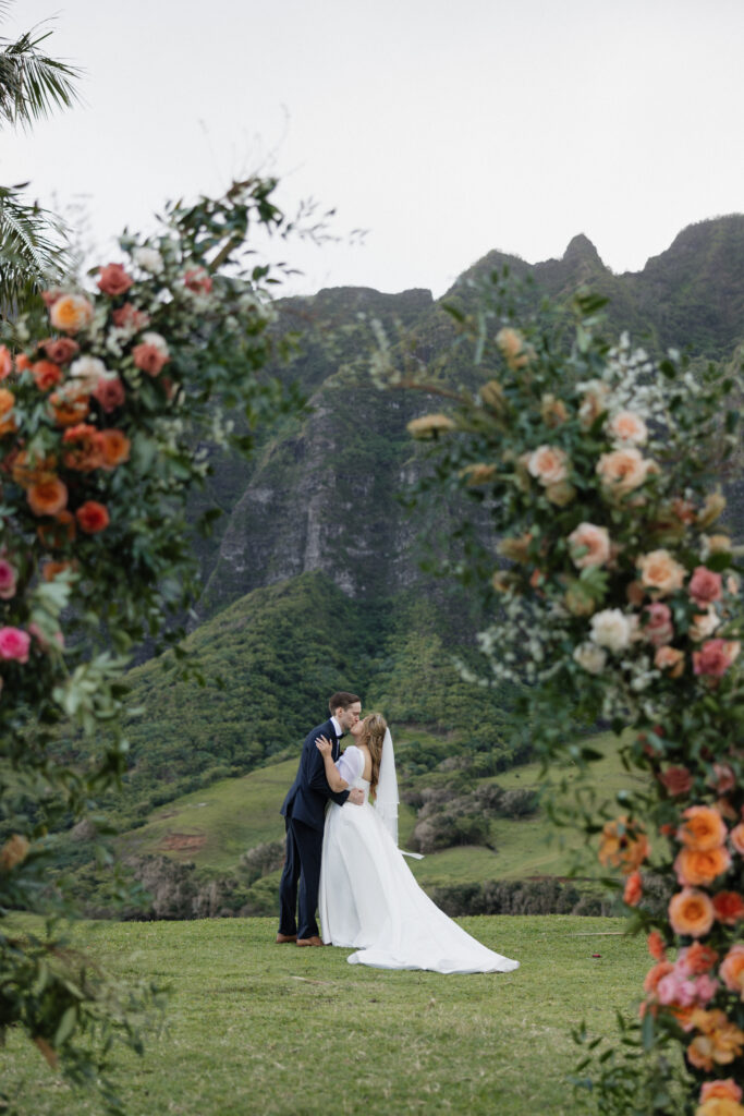 Hawaii wedding photographer captures bride and groom in frame between a large floral bouquet with cliffs behind them.