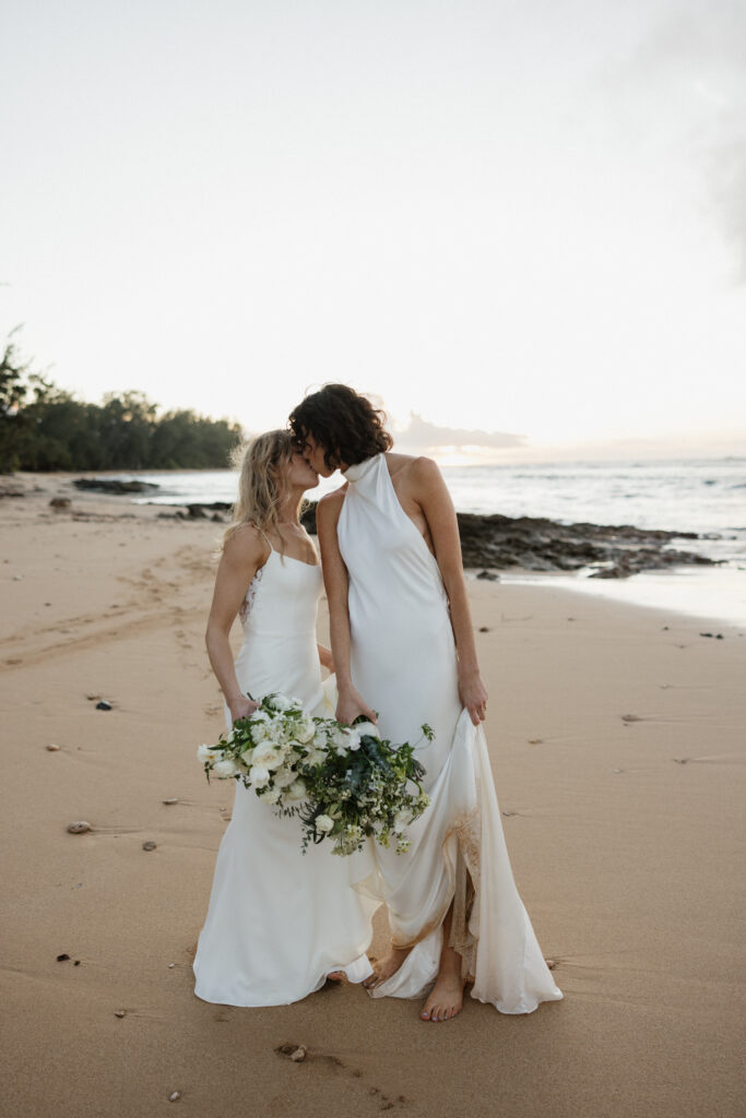 Brides stand on a sandy beach and kiss as the ocean is behind them during Hawaii wedding portraits. 