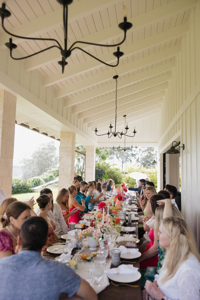 Guests gather around a long wooden table with colorful flower bouquets. The table is outside under a covered patio. 