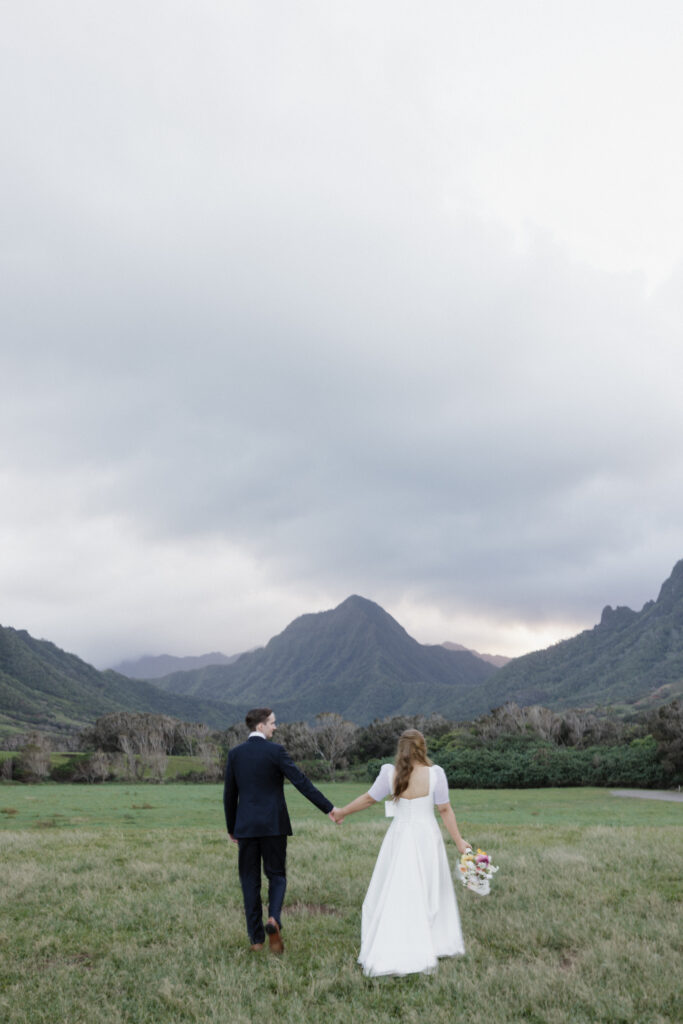 Bride and Groom hold their hands out as they walk through a grassy field with mountains behind them for their wedding portraits in Hawaii. 