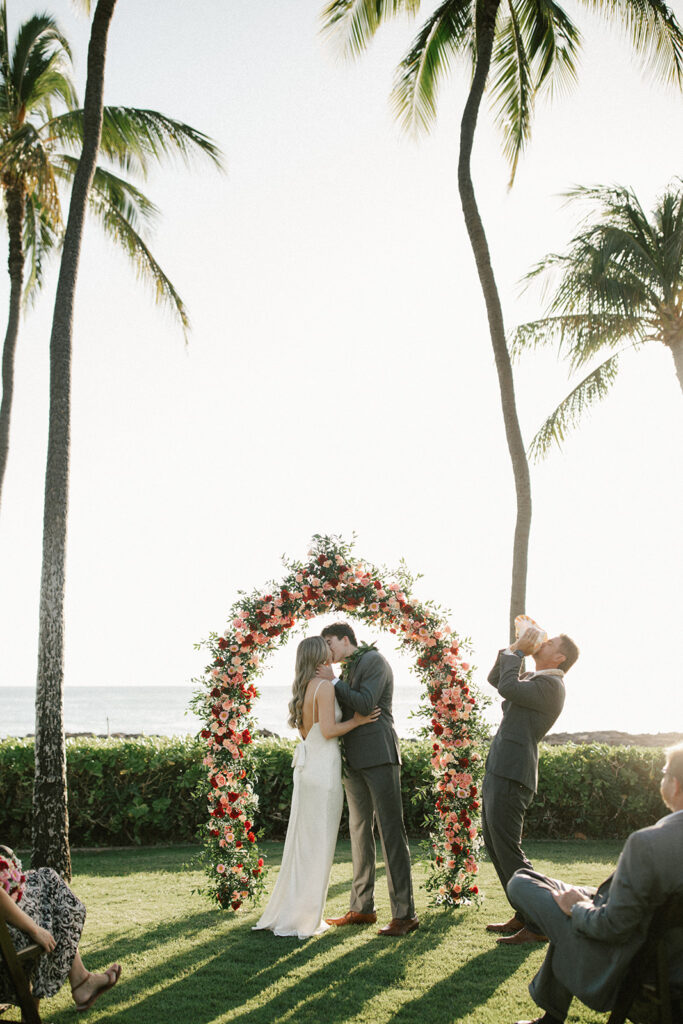 Bride and Groom kiss n front of colorful flower arch with palm trees around them as their groomsmen blows into a conch shell. 