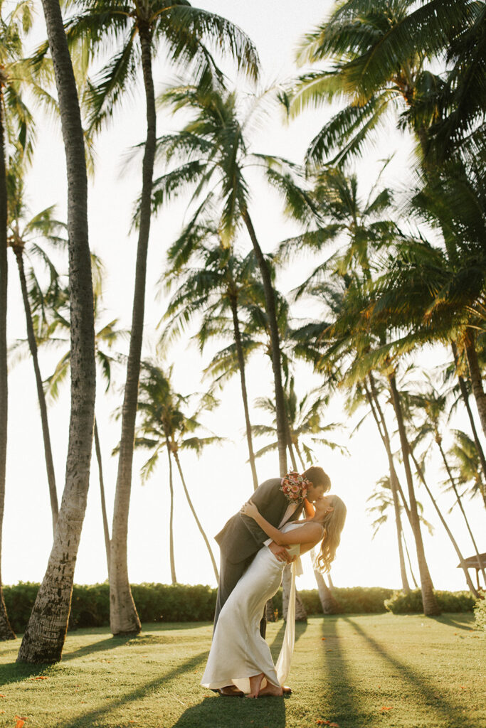 Groom dips bride for a kiss at sunset with palm trees swaying above them. 