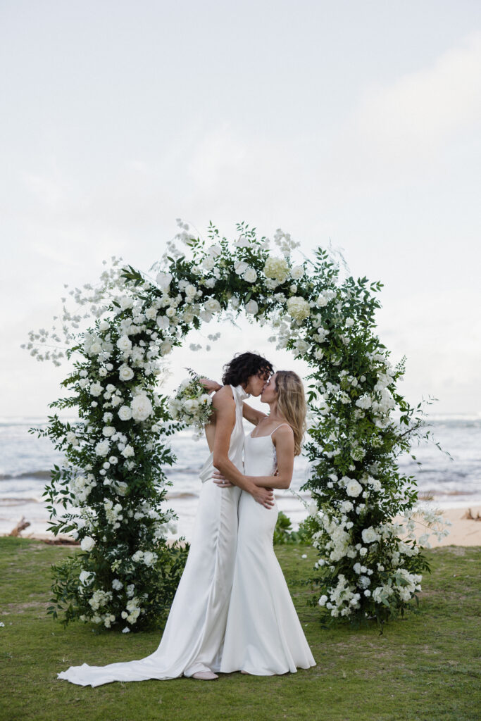 Brides, both in white dresses stand and kiss in front of a white floral arch on a beach front in Hawaii. 