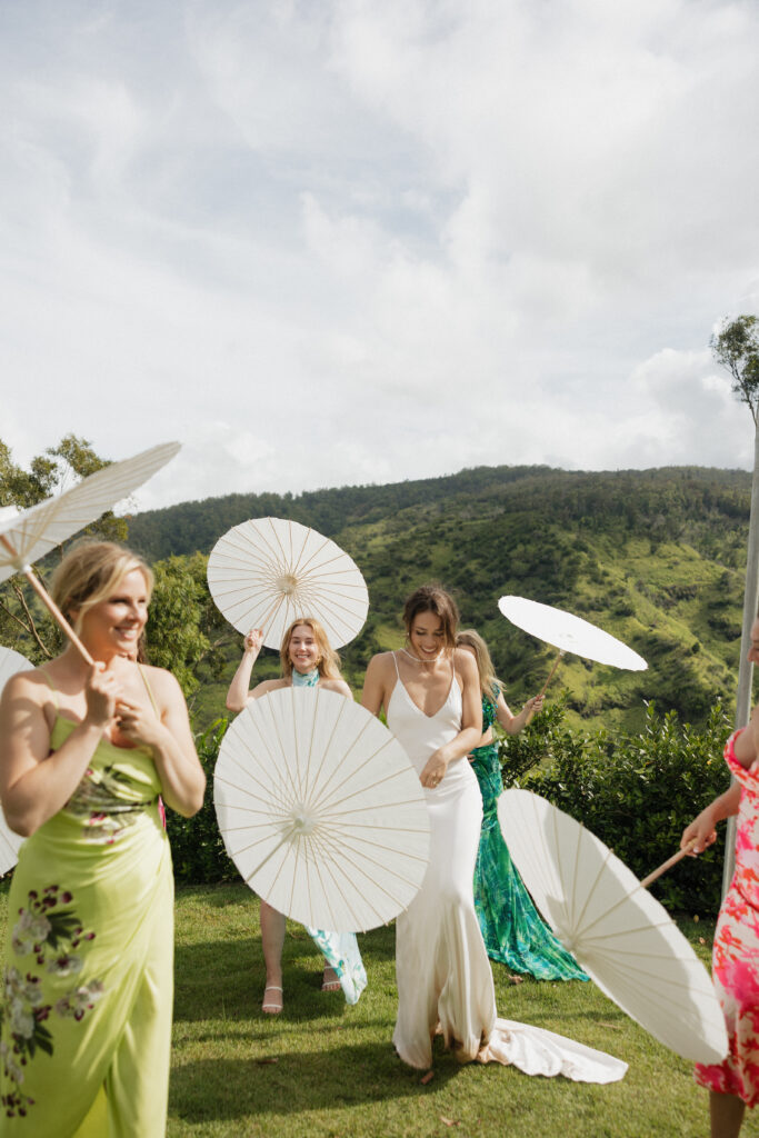 Bride stands center holding a white parasol umbrella as her bridesmaids walk around her holding white parasol umbrellas. 