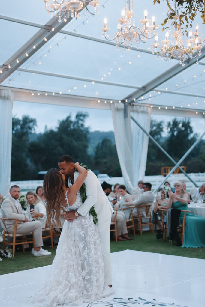 Groom dips bride for a kiss as they dance in their clear reception tent and guests watch lovingly. 