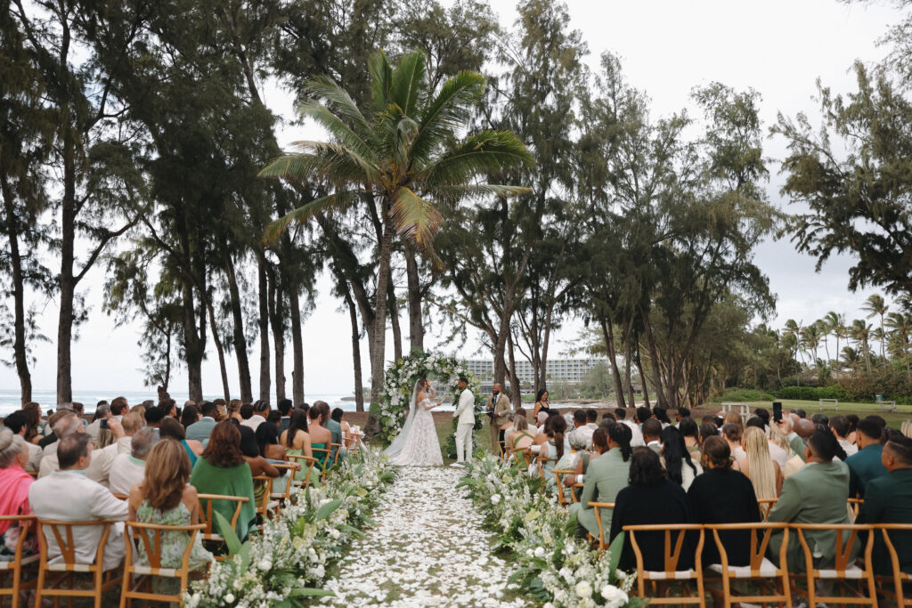 Bride and Groom stand at the end of the aisle with large green and white bouquets lining the aisle. Guests are seated in wooden chairs on both sides and white flower petals are scattered on the aisle. In the background is palm trees and Turtle Bay Resort. 