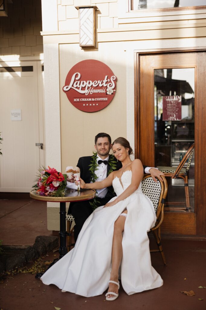 A bride and groom sit together outside of Lappert's Hawaii on Kauai eating ice cream on their elopement day. The brides bouquet of bright pink flowers sits on the table outside of the ice cream shop.