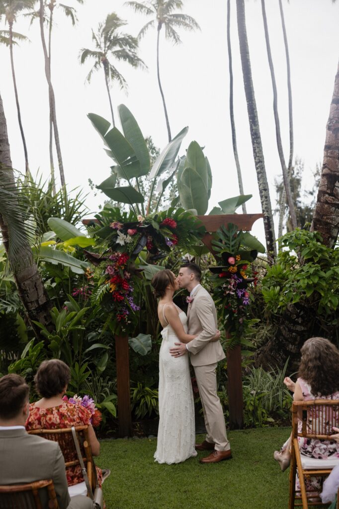 A couple kisses in front of a wooden arch covered in deep colored tropical flowers at their outdoor wedding ceremony at Male'ana Gardens in Hawaii.  